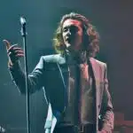 A formally dressed man confidently holds a microphone, possibly discussing the song "Love of My Life" and its connection to Harry Styles.