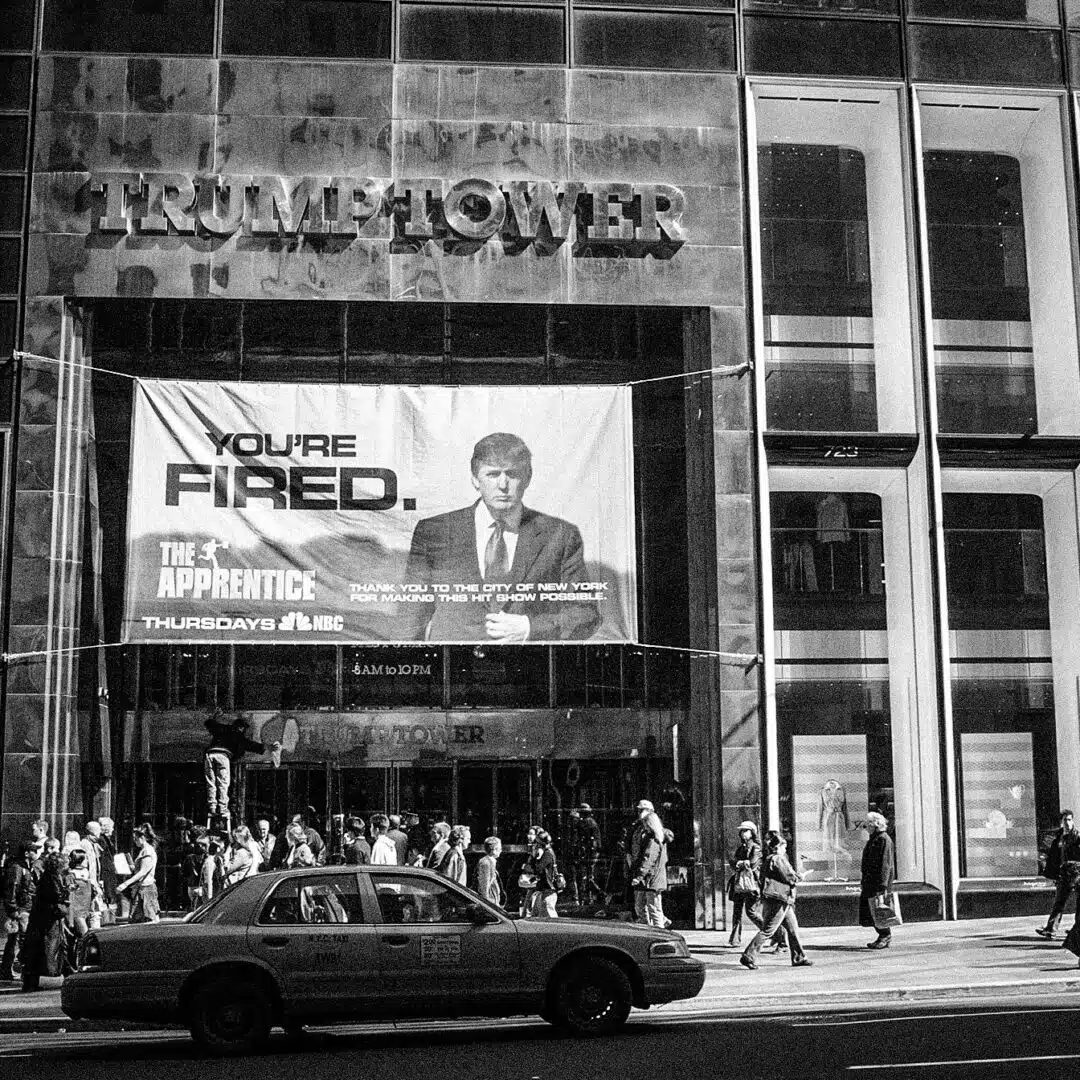 A sign advertising the television show is displayed on Trump Tower. The sign features a serious-looking Donald Trump walking and wearing a suit, with the words "You're Fired" in bold black lettering against a light blue background.