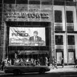 A sign advertising the television show is displayed on Trump Tower. The sign features a serious-looking Donald Trump walking and wearing a suit, with the words "You're Fired" in bold black lettering against a light blue background.